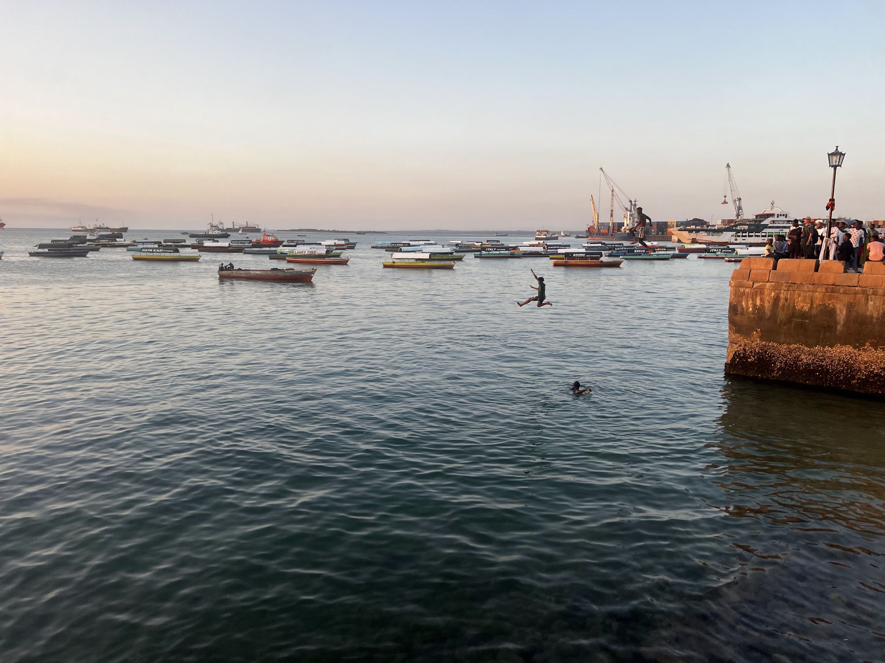 Boys jumping off a harbour wall at dusk in Stone Town, fishing boats behind